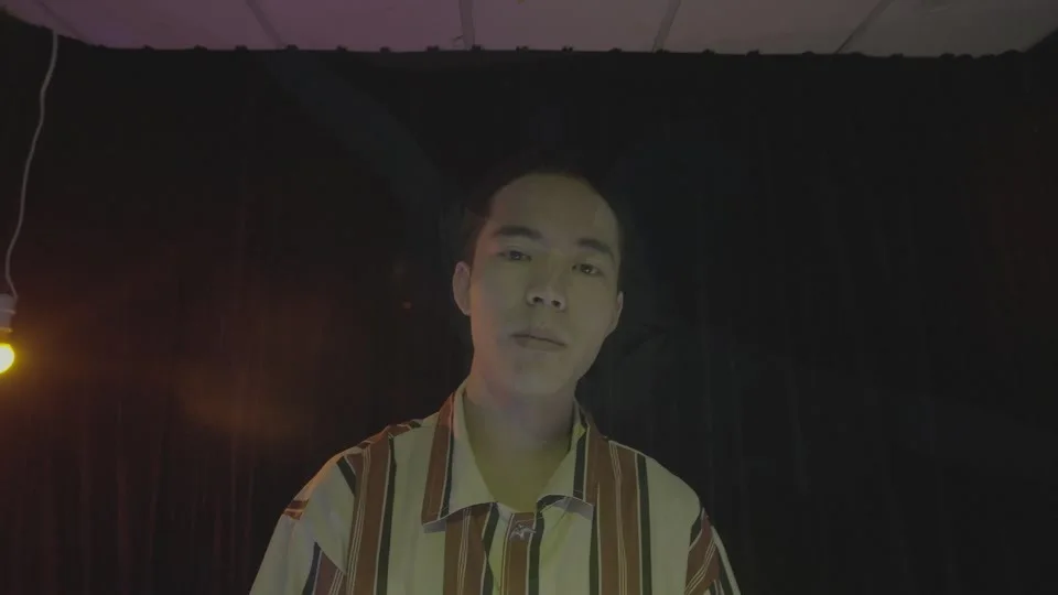 A performer in a striped shirt faces the camera beneath a hanging yellow bulb against a dark curtain in Shoot Self with You / Human Surrender.