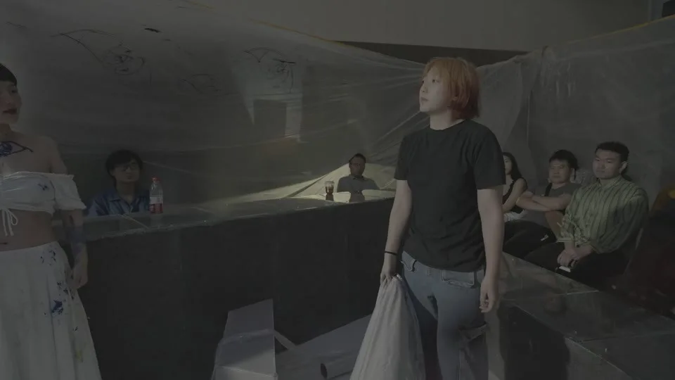 A standing figure holds white fabric while seated spectators remain visible behind translucent material in the public performance space.