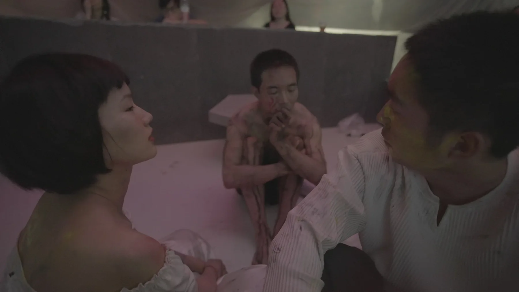 Three performers share a pink-lit room, with two figures facing each other in the foreground and a third seated between them in the background.