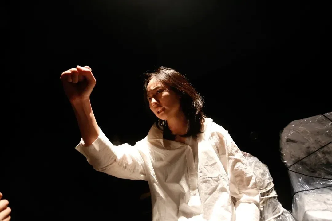 A performer in a loose white costume raises a fist under a spotlight against a dark stage.