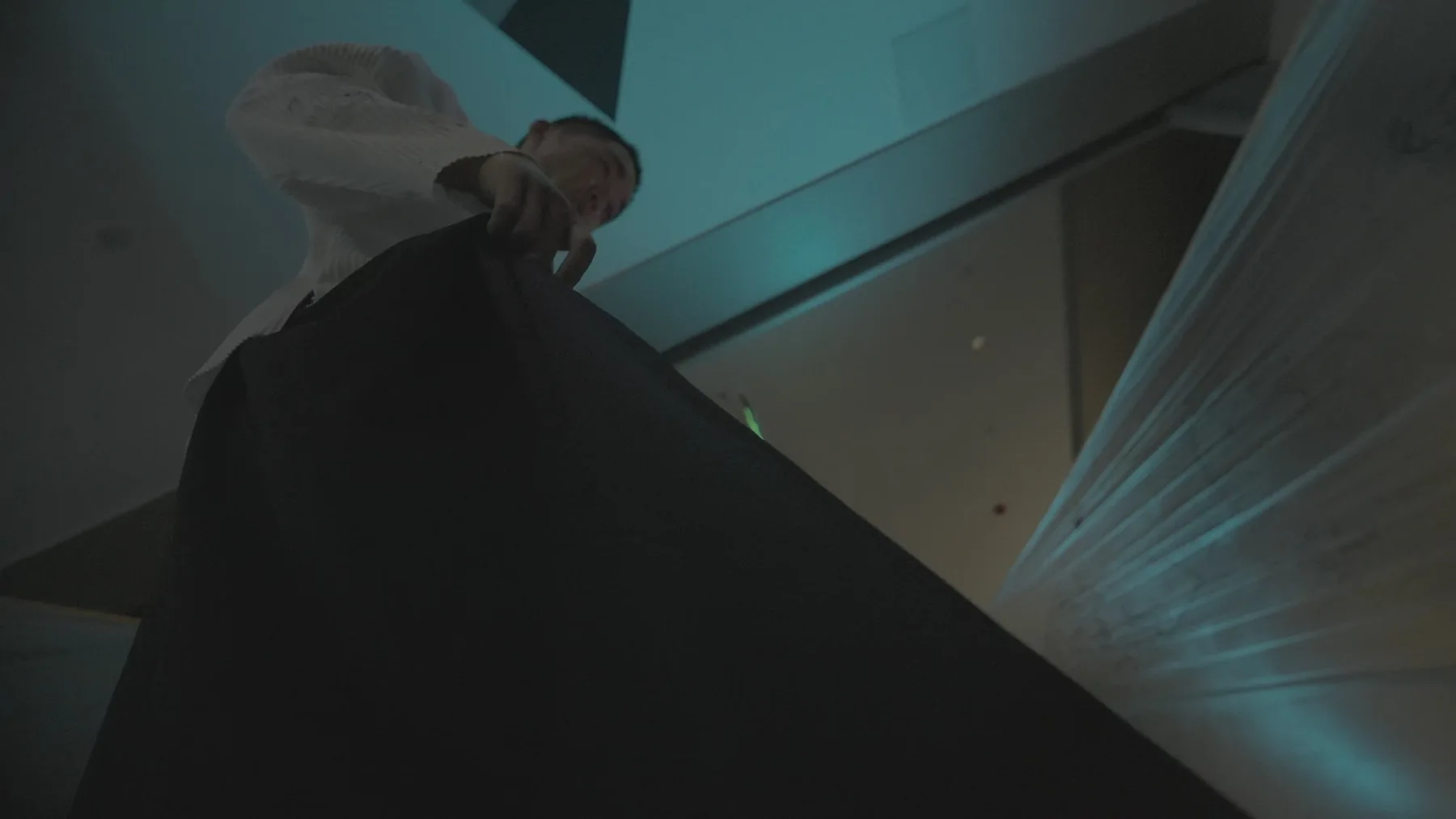 A performer seen from a low camera angle, with white fabric and reflective surfaces crossing the frame.