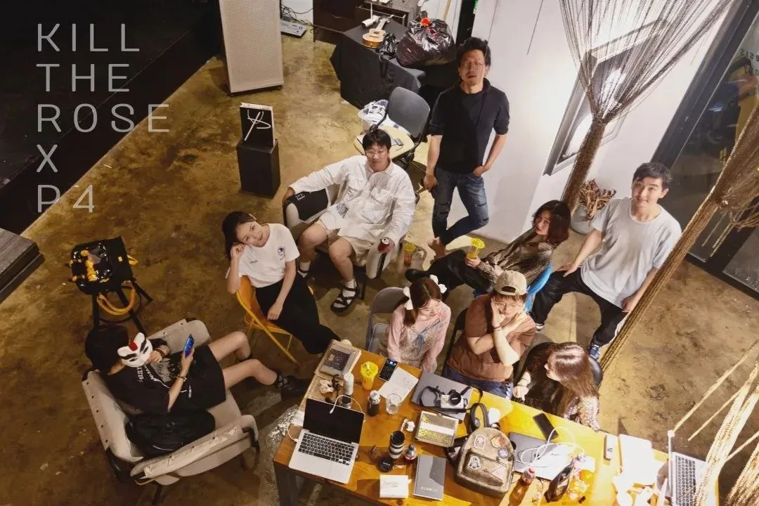 A group gathers around tables with laptops, cameras, drinks, and bags in an overhead project image marked “KILL THE ROSE X P4.”